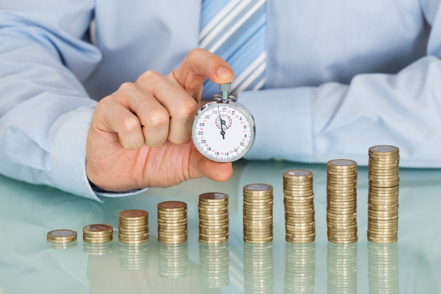 A businessperson holding a stopwatch behind ascending stacks of coins.